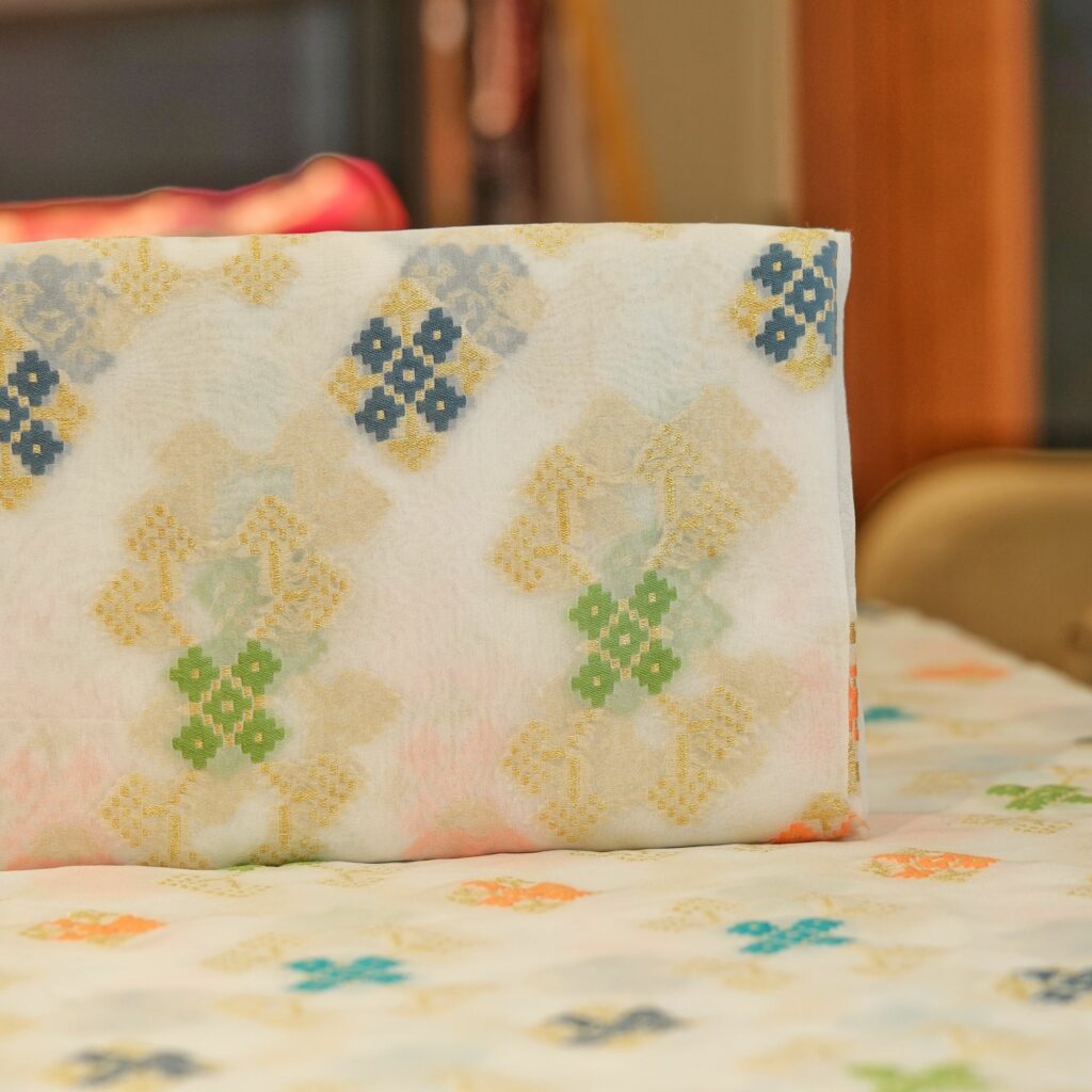 Sheer chiffon fabric with multicolored ikkat-style cotton floral motifs and shimmering golden zari weave on an off-white base.