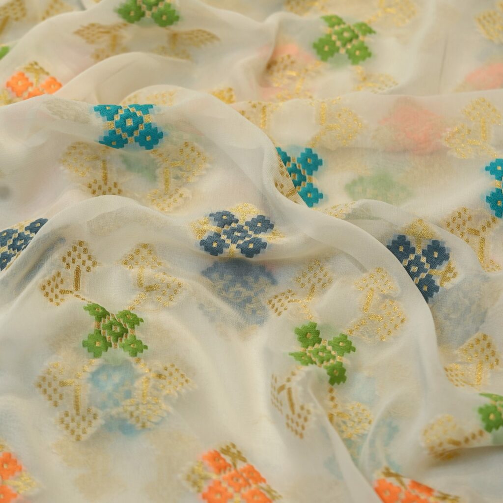 Sheer chiffon fabric with multicolored ikkat-style cotton floral motifs and shimmering golden zari weave on an off-white base.