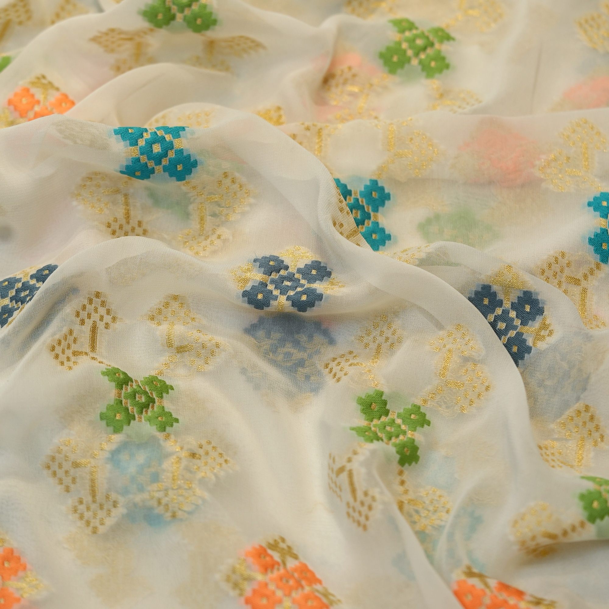 Sheer chiffon fabric with multicolored ikkat-style cotton floral motifs and shimmering golden zari weave on an off-white base.